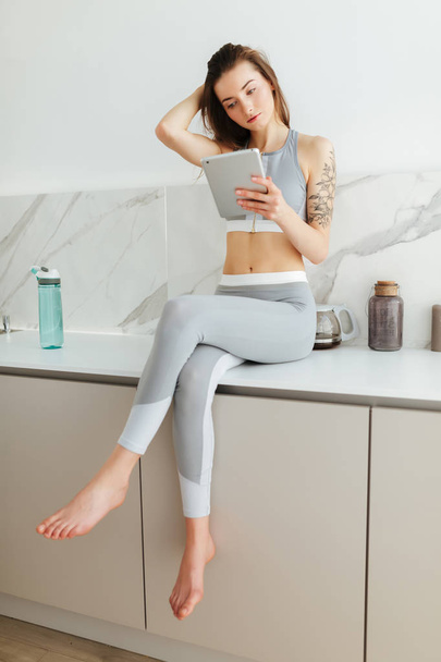 Beautiful girl in sporty top and leggings sitting on kitchen table with laptop in hand and thoughtfully looking aside isolated - Photo, Image
