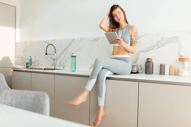 Pretty lady in sporty top and leggings sitting on kitchen table with tablet in hand and thoughtfully looking aside isolated - Photo, Image