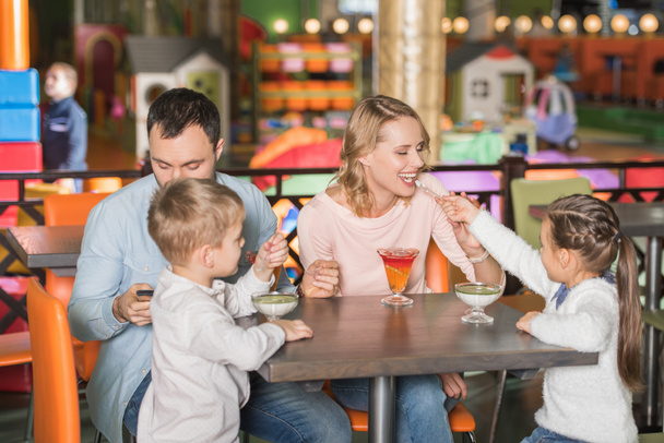 οικογένεια με δύο παιδιά τρώγοντας νόστιμα επιδόρπια και πατέρας του χρησιμοποιώντας το smartphone στο καφέ - Φωτογραφία, εικόνα