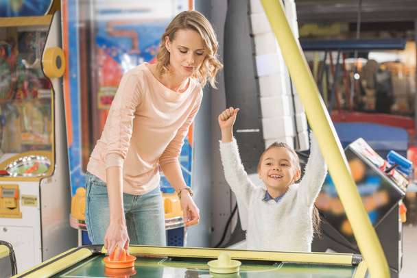 smiling mother and triumphing little daughter playing air hockey in game center - Photo, Image