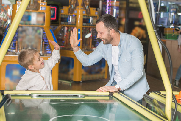 onnellinen isä ja poika antavat yläfemmat pelatessaan ilmakiekkoa viihdekeskuksessa
 - Valokuva, kuva