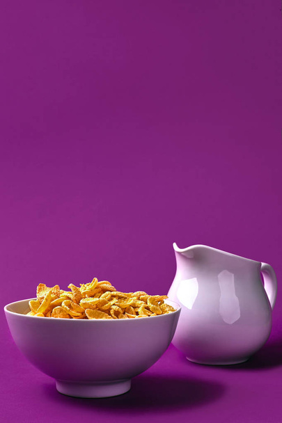 Cuenco con hojuelas de maíz, jarra de leche sobre fondo morado. oncept de un desayuno saludable
 - Foto, Imagen