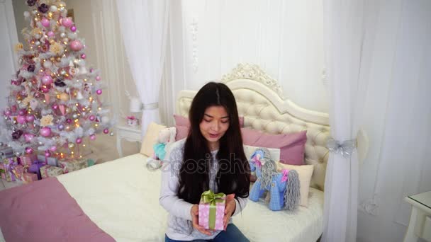 Belle fille dans l'humeur du Nouvel An avec un cadeau à la main souriant à la caméra et assis sur le lit en arrière-plan de l'arbre de Noël dans la chambre rose
. - Séquence, vidéo
