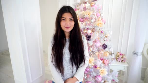 Δελεαστικός κορίτσι σε αναμονή για το νέο έτος γιορτές χαμογελά στη φωτογραφική μηχανή σε φόντο διακοσμημένο χριστουγεννιάτικο δέντρο σε λευκό δωμάτιο. - Πλάνα, βίντεο