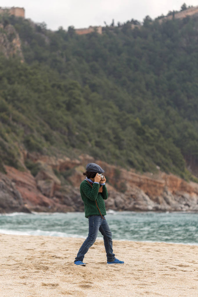 Teenager boy wearing blue jeans, dark green sweater and flat cap photographing at the beach. Vertical composition, shallow depth of field. - Foto, immagini