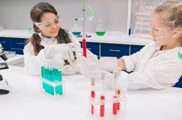 Two little kids in lab coat learning chemistry in school laboratory. Young scientists in protective glasses making experiment in lab or chemical cabinet. Studying ingredients for experiments . - Foto, Bild