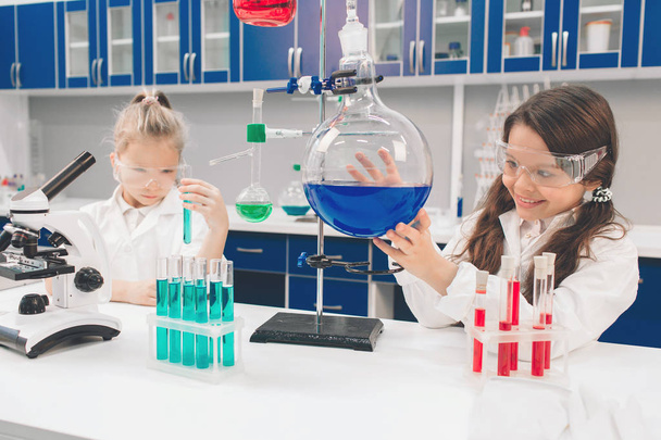 Two little kids in lab coat learning chemistry in school laboratory. Young scientists in protective glasses making experiment in lab or chemical cabinet. Studying ingredients for experiments . - Zdjęcie, obraz