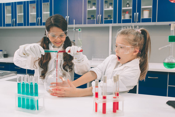 Zwei kleine Kinder im Laborkittel lernen Chemie im Schullabor. Junge Wissenschaftler in Schutzbrillen experimentieren im Labor oder Chemiekabinett. Inhaltsstoffe für Experimente studieren . - Foto, Bild