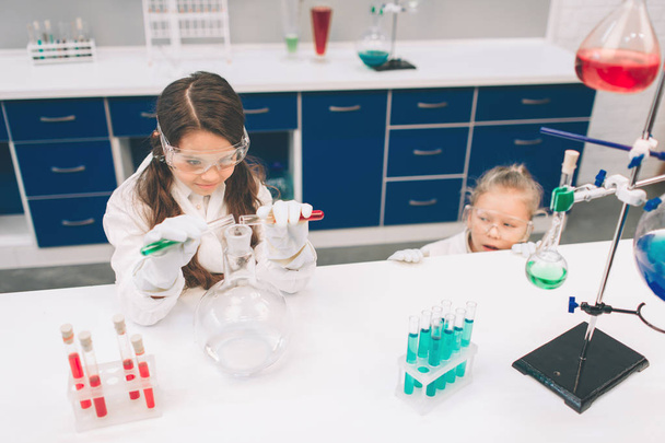 Two little kids in lab coat learning chemistry in school laboratory. Young scientists in protective glasses making experiment in lab or chemical cabinet. Studying ingredients for experiments . - Foto, afbeelding