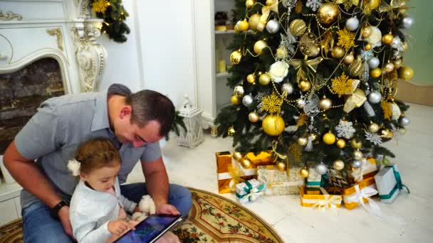 Buen padre y niña pasan tiempo y sosteniendo el gadget sentado en el piso en una habitación grande en el fondo del árbol de Navidad decorado y la chimenea
. - Metraje, vídeo