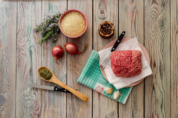 Cooking food. Raw, fresh minced meat on a plate, spices, rice and vegetables on a wooden table close-up. - Photo, Image