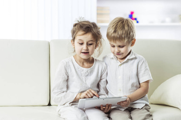 младшие брат и сестра с цифровым планшетом сидит на диване в детской
. - Фото, изображение
