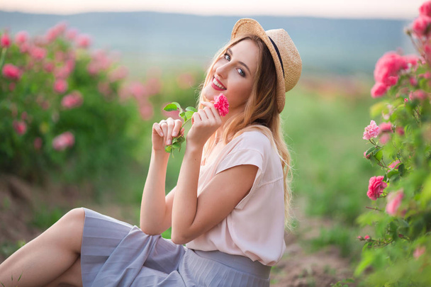 Beautiful young girl is wearing hat having rest in a garden with pink blossom roses - 写真・画像