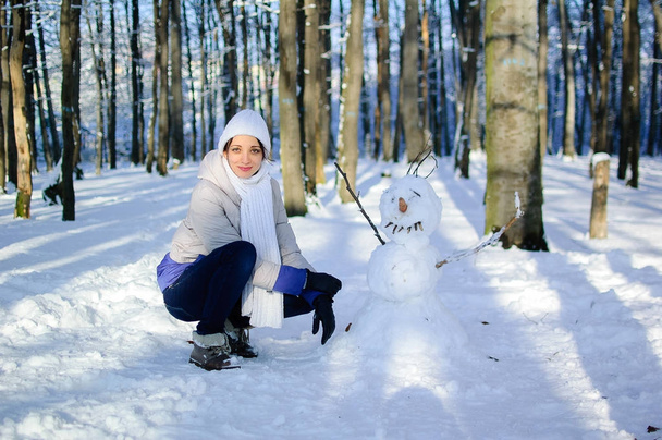 Retrato de una chica con sombrero blanco y bufanda afuera sentada cerca de un muñeco de nieve divertido. Cielo azul y árboles helados en el fondo
. - Foto, Imagen