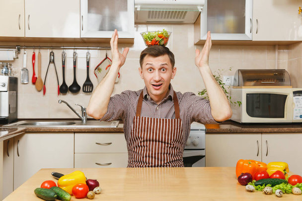 Vonzó szórakoztató kaukázusi fiatalember, kötény, barna inget növényi saláta tál feje világos konyha asztalnál ül. Diéta fogalma. Egészséges életmód. Főzés otthon. Készítsünk élelmiszer. - Fotó, kép