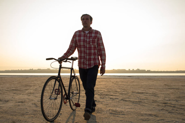 male bicycle rider walking on the sand, sunrise and lake or river background - Φωτογραφία, εικόνα