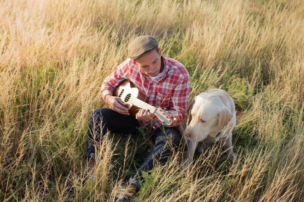 Young hipster and golden labrador dog having good time in the fields and play on ukulele - Fotó, kép