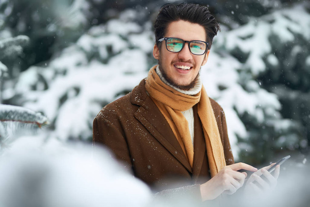 Close-up portrait of young beautiful men. Smiling young man having fun outdoors.the man is using the phone. A man loves winter. He is wearing a coat, a sweater and a scarf. Winter concept. Snowfall. - Foto, Imagem