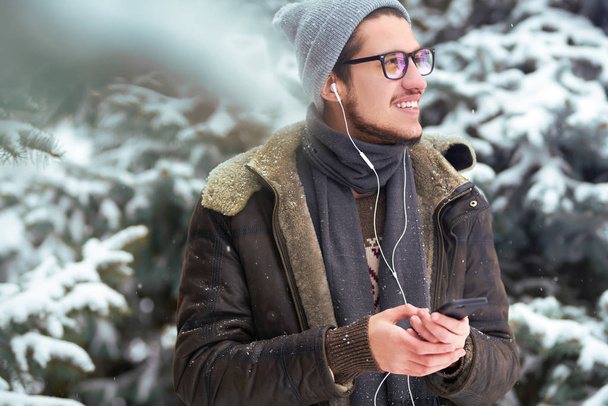 Close-up portrait of young beautiful men. Smiling young man having fun outdoors.the man is using the phone. A man loves winter. He is wearing a coat, a sweater and a scarf. Winter concept. Snowfall. - Foto, Imagen