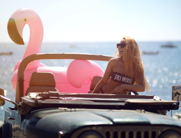young girl in sunglasses sitting in beach truck car on sunny summer day near sea beach with giant inflatable giant pink flamingo float mattress - Photo, Image