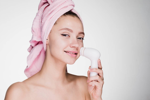 Gelukkige vrouw met een handdoek op haar hoofd reinigt de huid van haar gezicht met een borstel voor diepe reiniging - Foto, afbeelding