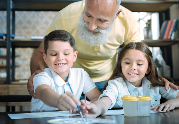 Careful grandfather looking at kids painting together - Foto, imagen
