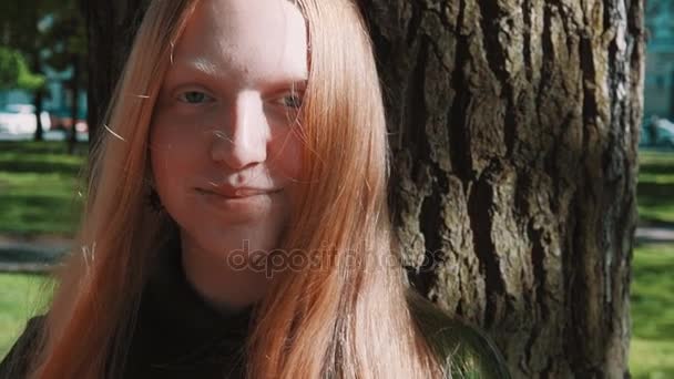 Longhaired redhead young girl standing under park tree - Filmagem, Vídeo