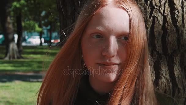 Dlouhovlasého zrzka hezká dívka usmívá pod stromem v parku - Záběry, video