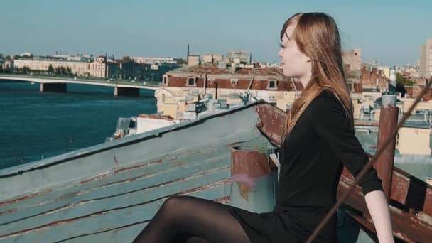 attraktive junge Frau im schwarzen Kleid sitzt auf einem Dach mit malerischem Stadtbild - Filmmaterial, Video