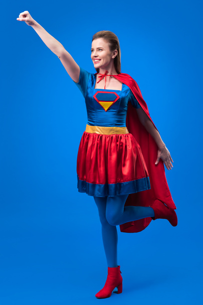 mujer sonriente en traje de superhéroe con brazo extendido aislado en azul
 - Foto, Imagen