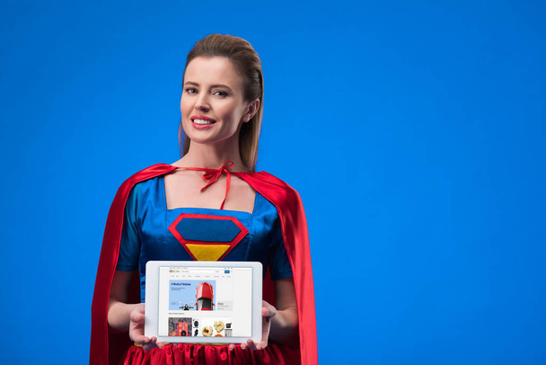 retrato de mujer sonriente en traje de superhéroe mostrando tableta aislada en azul
 - Foto, Imagen