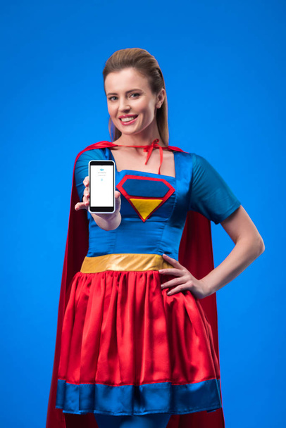 πορτρέτο του χαμογελαστό γυναίκα στο κοστούμι superhero δείχνει smartphone που απομονώνονται σε μπλε - Φωτογραφία, εικόνα