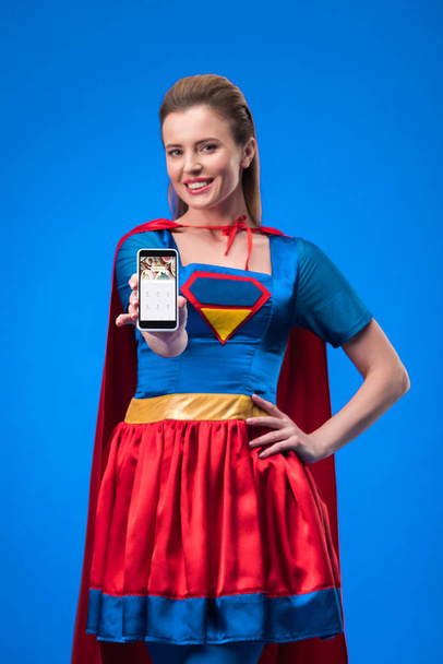 portrait of smiling woman in superhero costume showing smartphone isolated on blue - Fotó, kép