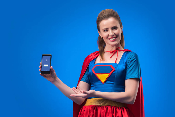 Muotokuva hymyilevä nainen supersankari puku osoittaa älypuhelin facebook app eristetty sininen
 - Valokuva, kuva
