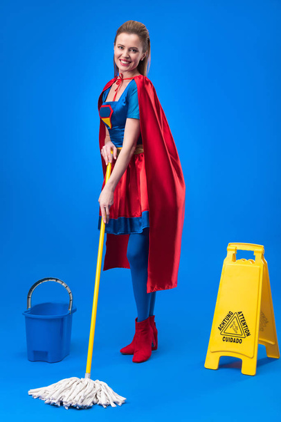 femme souriante en costume de super-héros avec signe de prudence, seau et balai pour le nettoyage isolé sur bleu
 - Photo, image