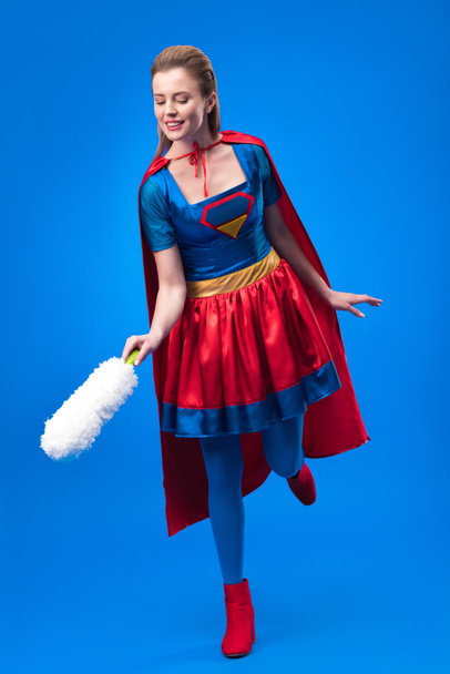 Lachende vrouw in superheld kostuum met stof Reinigingsborsteltje geïsoleerd op blauw - Foto, afbeelding