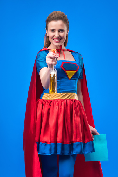smiling woman in superhero costume with rag and detergent in hands isolated on blue - Foto, afbeelding