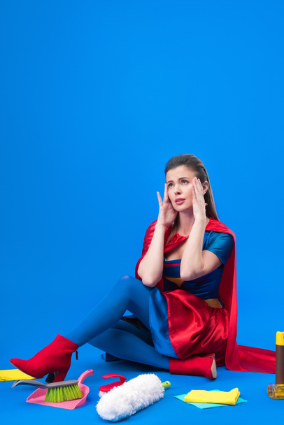 thoughtful woman in superhero costume with cleaning supplies around isolated on blue - Фото, зображення