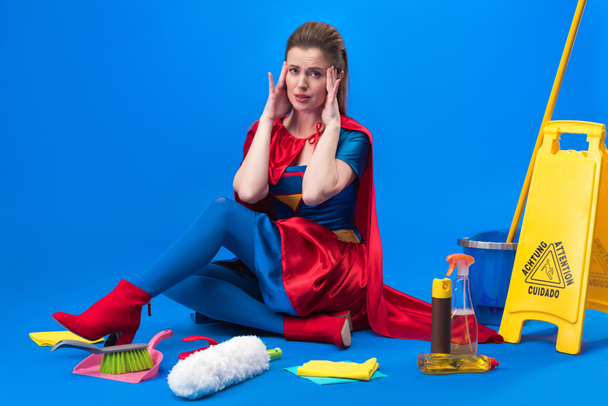 tired woman in superhero costume with cleaning supplies around isolated on blue - Fotó, kép