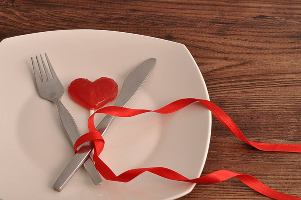 A fork an knife tied together with a red ribbon displayed on a white plate with a red heart shape lollipop - Fotó, kép