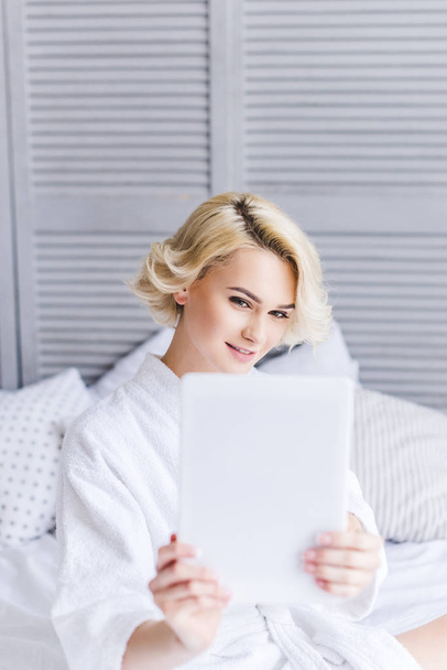 selective focus of smiling young woman in bathrobe using digital tablet in bedroom - Photo, Image