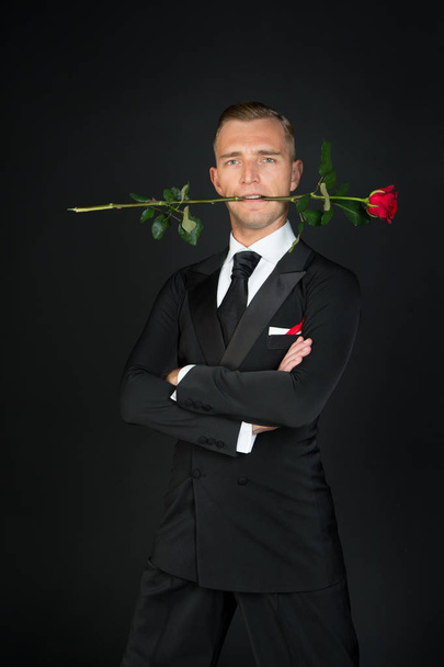 Man with red rose in mouth on dark background. Ballroom dancer in suit with flower. Valentines day concept. Love, romance, desire, proposal, date. Holiday celebration, party. - 写真・画像