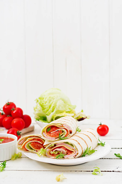 Tortilla kääri kinkkua, juustoa ja tomaatteja valkoisella puupohjalla
 - Valokuva, kuva