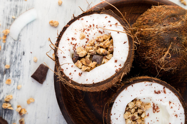 Здоровый завтрак с кокосовым бантом на белом фоне. Йогурт в кокосовой миске с кокосовыми хлопьями, шоколадом и мюсли. Вид сверху, плоский, накладные расходы
 - Фото, изображение