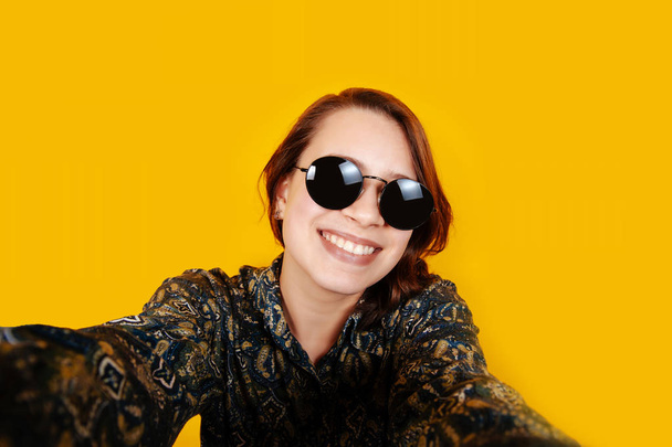 Allegro felice giovane donna con occhiali da sole guardando la fotocamera e prendendo selfie su sfondo arancione in studio. Sorridente ragazza felice
. - Foto, immagini