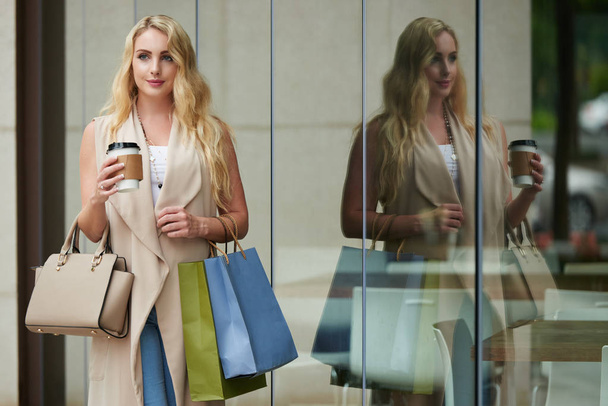 Portret van lachende blonde vrouw verlaten winkelcentrum met tassen en kopje koffie - Foto, afbeelding