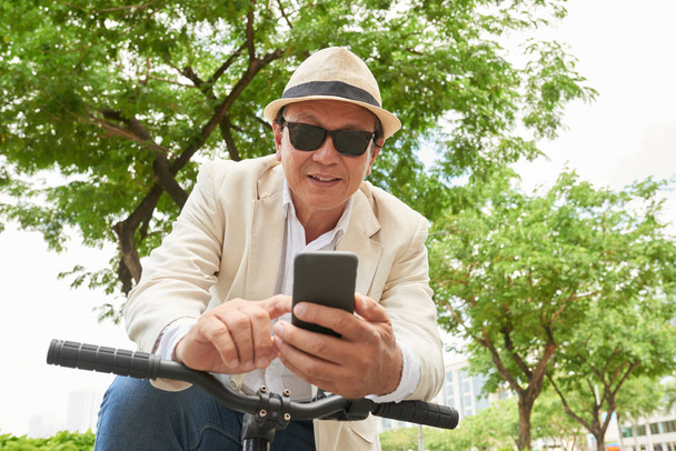 Lachende volwassen man leunend op zijn fiets en met behulp van smartphone - Foto, afbeelding