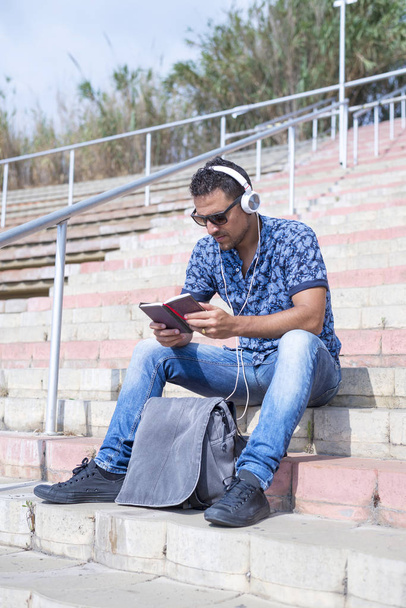 Happy attractive man listening music and typing phone sitting on stairs - Photo, image