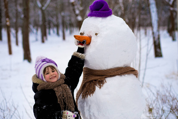 A small cheerful girl holds a big carrot, the nose of a big snowman. A cute little girl has fun in winter park, wintertime - Фото, изображение