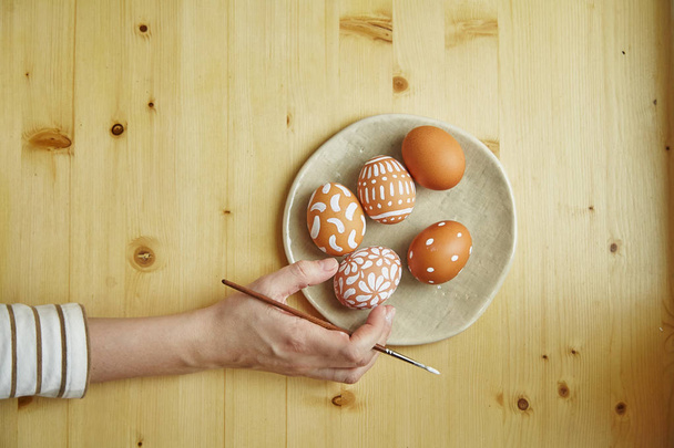 female hand holding brush over table with painted eggs with various white patterns for Easter holiday on plate     - Foto, Imagem
